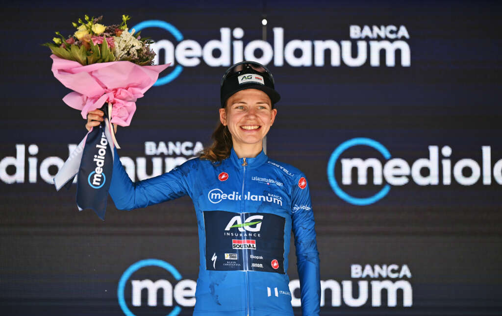 A young female cyclist wearing a blue jersey stands on stage holing up a bouquet of flowers