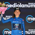 A young female cyclist wearing a blue jersey stands on stage holing up a bouquet of flowers