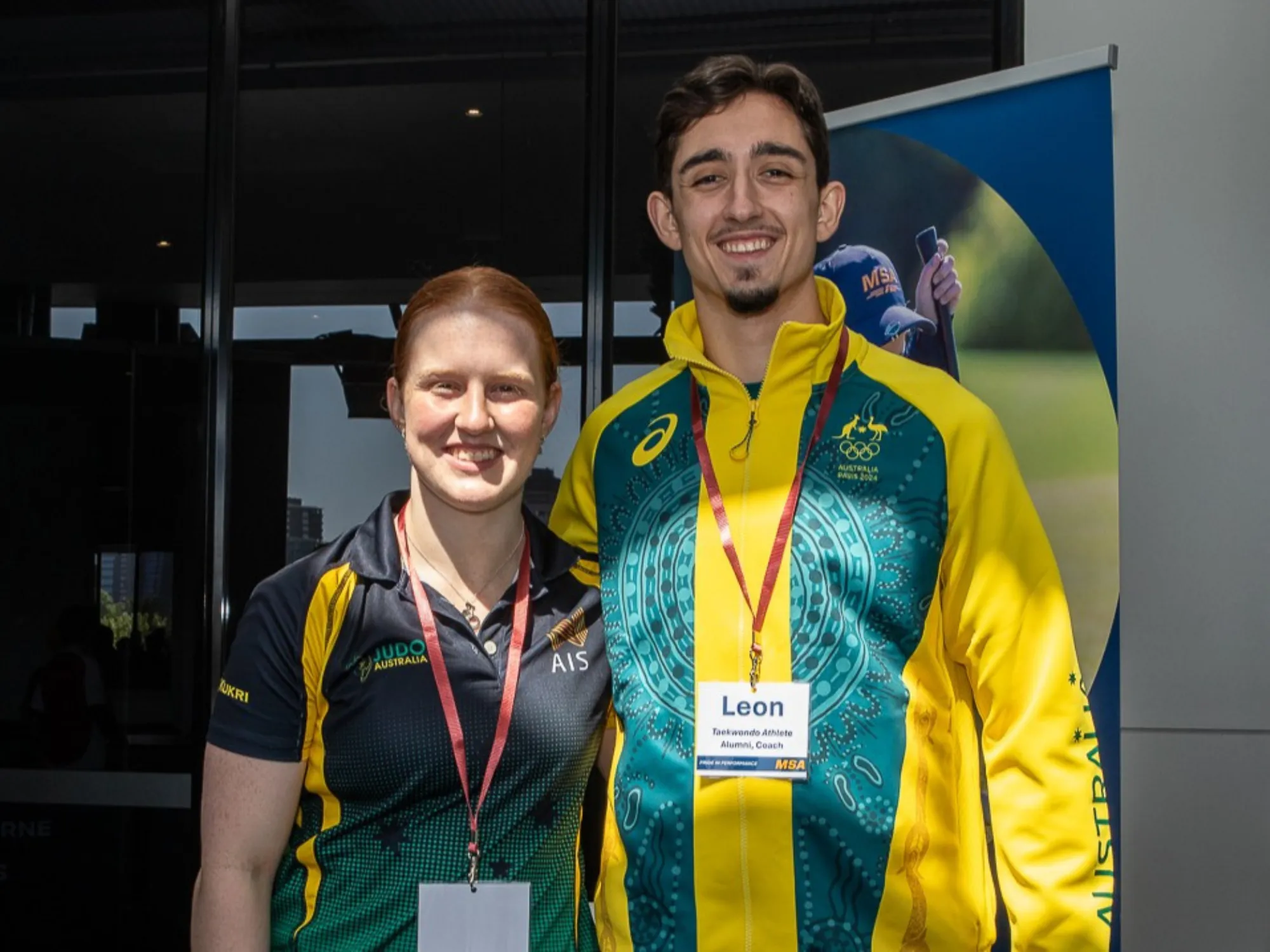 A female and male athlete both in their twenties standing side by side. The female (left) wears a Judo Australia polo with a name lanyard, the male (right) wears Australian Olympic team uniform with a name lanyard.