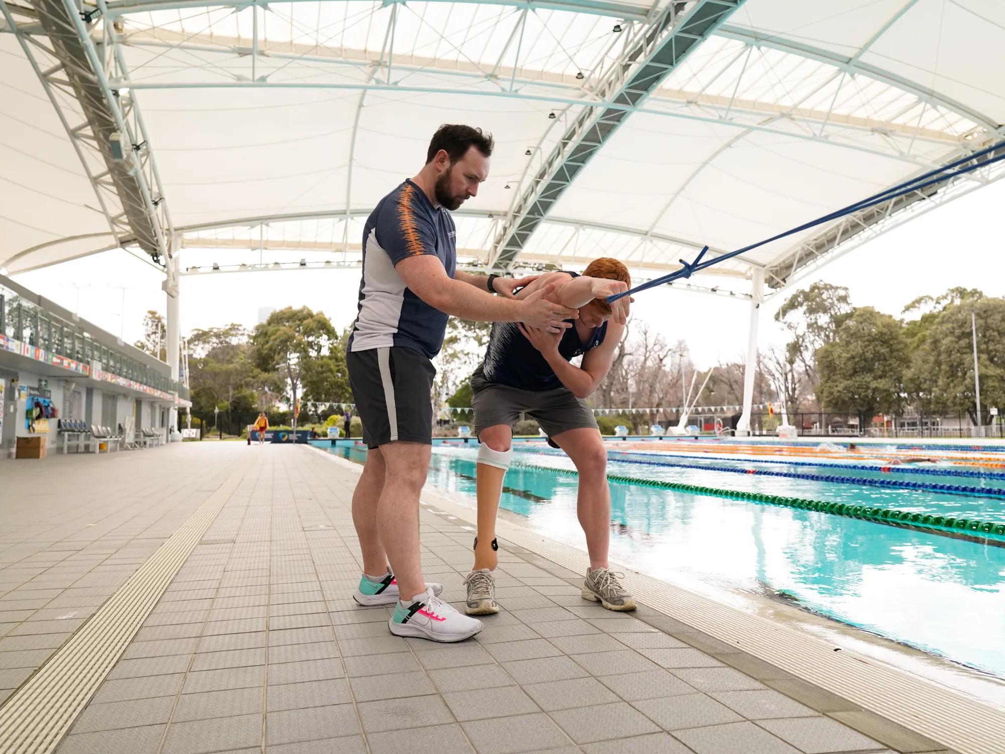 A brown haired man with a beard in VIS uniform is standing next to a red-haired athlete who is doing resistance band arm exercises.