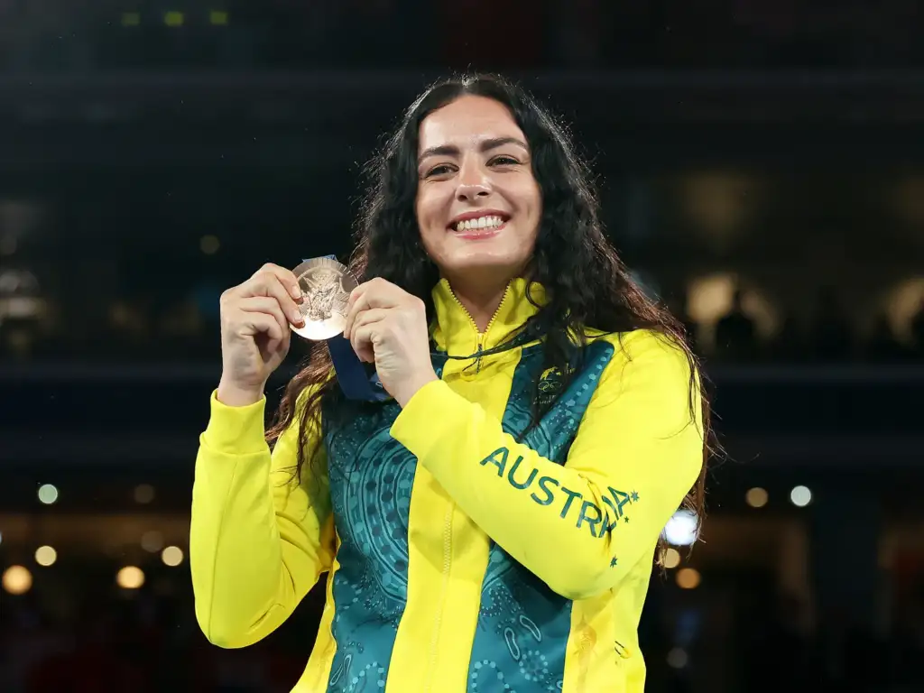 A photo of a Caucasian female with long dark brown wavy hair, wearing Australian Olympic Team uniform and holding up a bronze medal.
