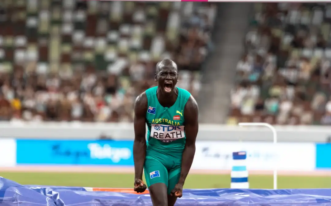 A male in his twenties is wearing green Australian Team uniform and is screaming with triumph after completing high jump. A high jump bar is in the background.