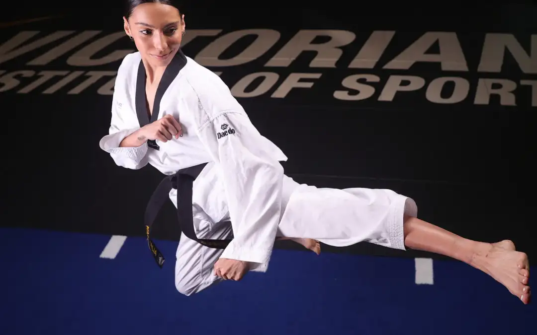 A female in her twenties with brown hair, wearing white taekwondo uniform, is mid-air performing a taekwondo kick.