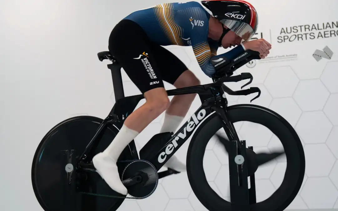 A young male athlete wearing VIS lyrca is riding on a stationary bike in a white room.