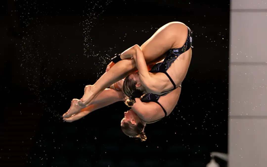 Two female athletes are wearing Australian Team uniform bathers and are mid-air whilst performing a diving trick.