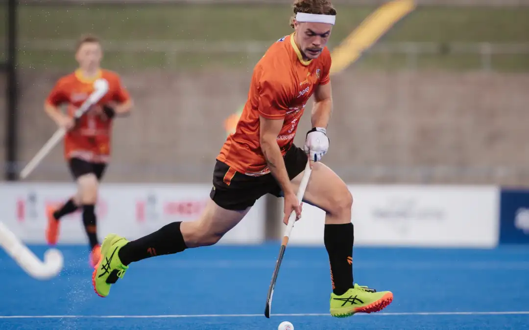 A male athlete in his twenties with brown hair and wearing an Australian Team uniform is running in action whilst dribbling a hockey ball.