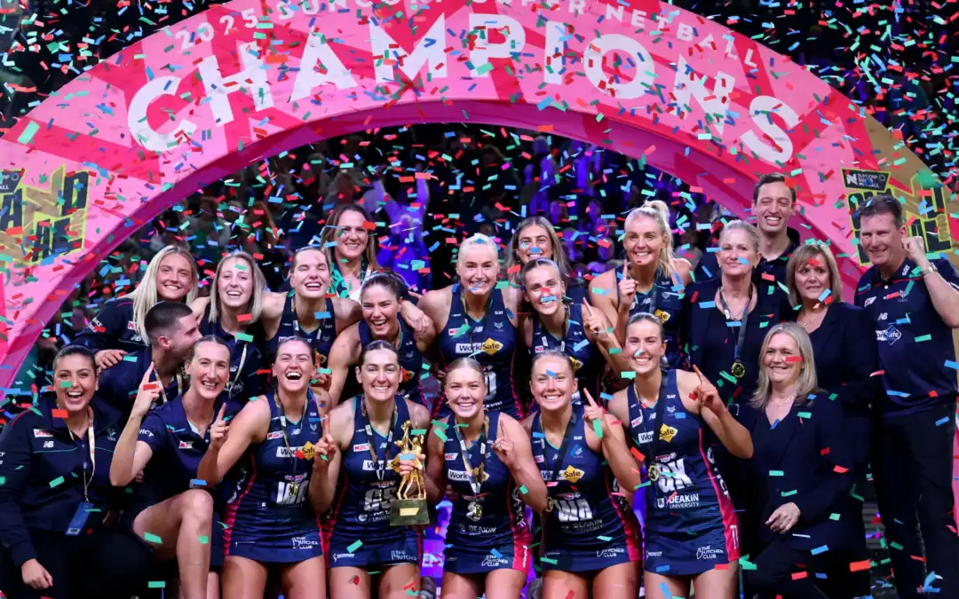 A group of female netballers and some support staff stand on a podium with confetti raining down, and a banner reading CHAMPIONS behind them.