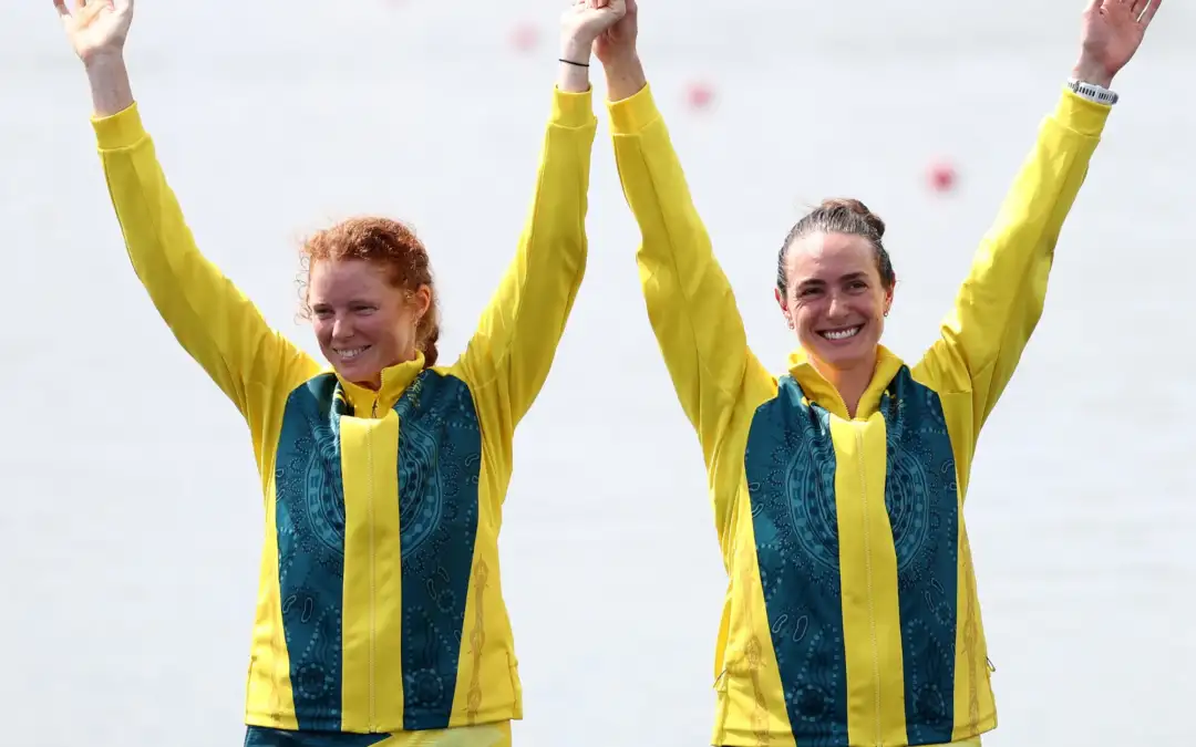 Two females in their 20s wear Australian Olympic Team uniform and stand holding hands with their arms in the air.