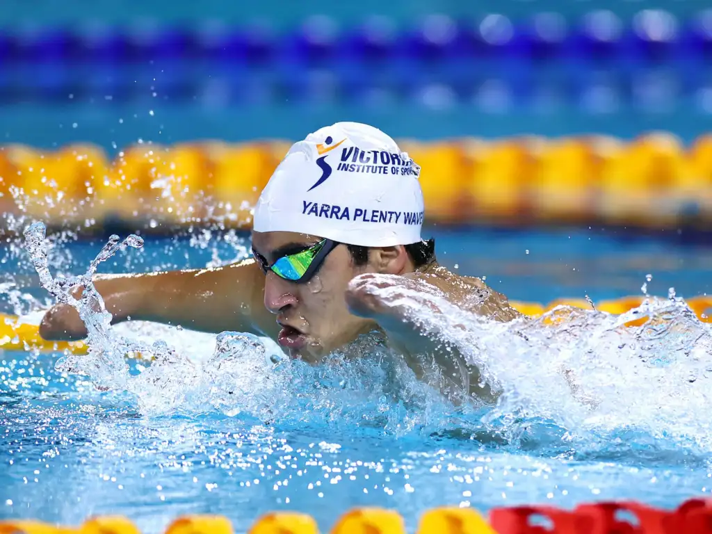 A male swimmer wearing a white VIS branded swimming cap is mid-stroke. He has a double arm and leg impairment.
