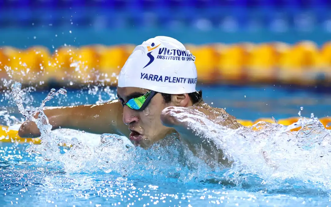 A male swimmer wearing a white VIS branded swimming cap is mid-stroke. He has a double arm and leg impairment.