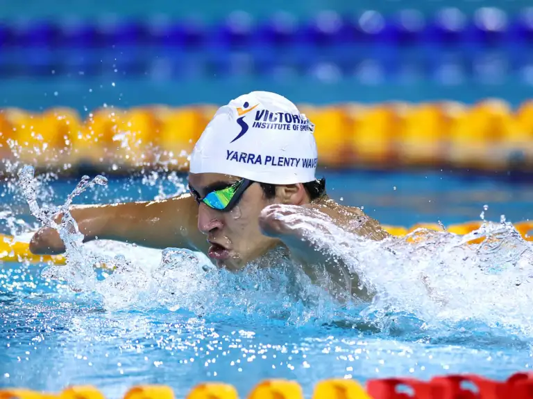 A male swimmer wearing a white VIS branded swimming cap is mid-stroke. He has a double arm and leg impairment.