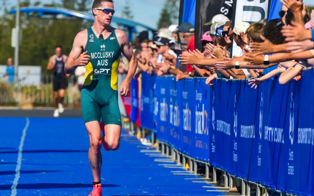 A male adult is wearing an Australian triathlon suit and running down a blue carpeted runway towards a finish line. Fans cheer him on on the right hand side.
