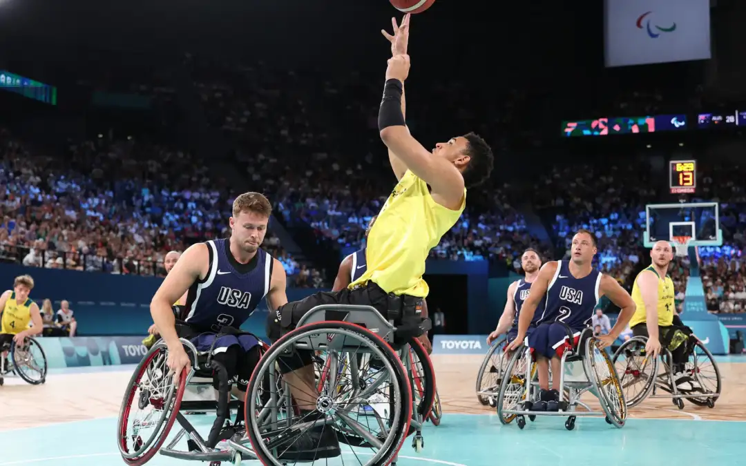 A male athlete in his twenties who is a wheelchair user is performing a basketball layup during a wheelchair basketball match.
