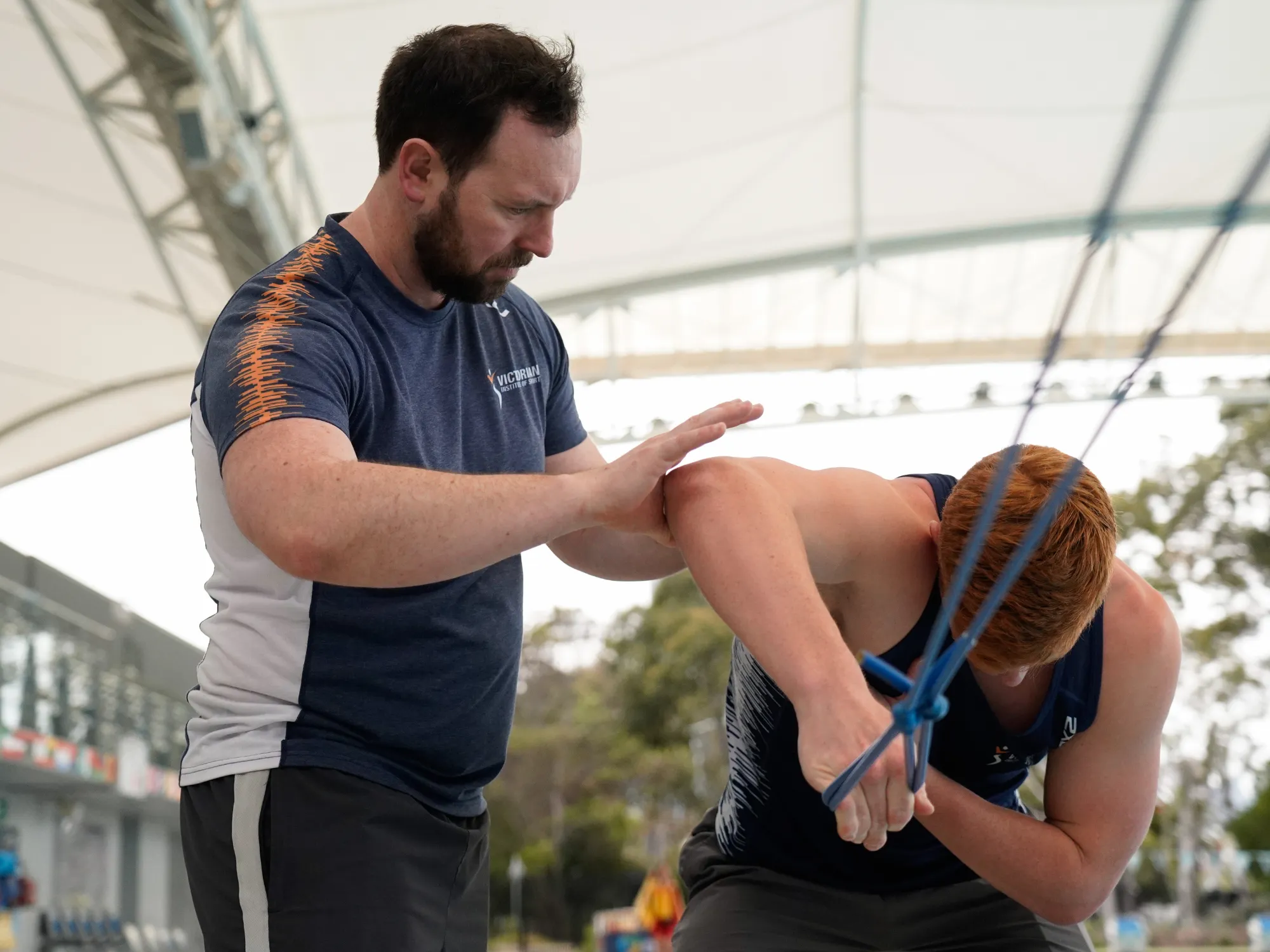 A brown haired man with a beard in VIS uniform is standing next to a red-haired athlete who is doing resistance band arm exercises. 
