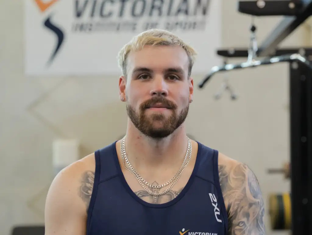 A headshot of a male athlete with short blonde hair and a dark brown beard. He stands in front of a blurred background with gym equipment and a sign reading Victorian Institute of Sport.