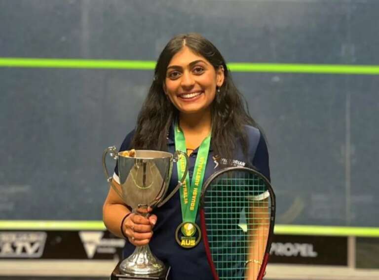 a headshot of a young female athlete with long wavy black hair. She is smiling to camera, wearing a navy polo, a medal around her neck with a green strap, and in her hands she holds a trophy cup and a squash racquet.