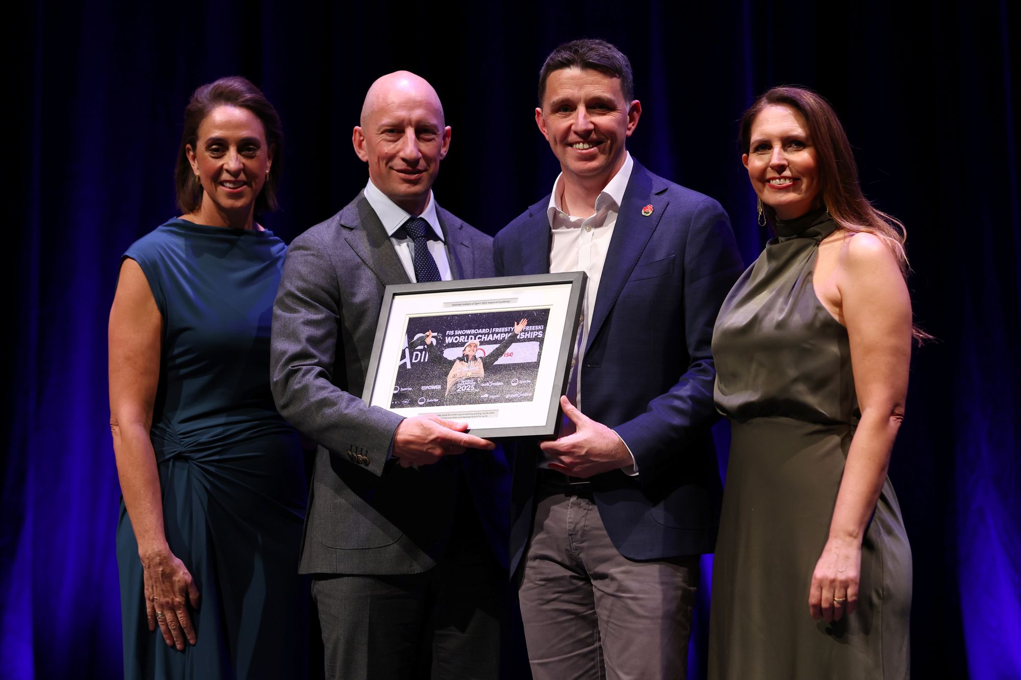 A photo of four adults standing on stage, all smiling to camera. From left to right is Nicole Livingstone AO, Will Morgan, Ryan Batchelor MP, Lauren Burns OAM.