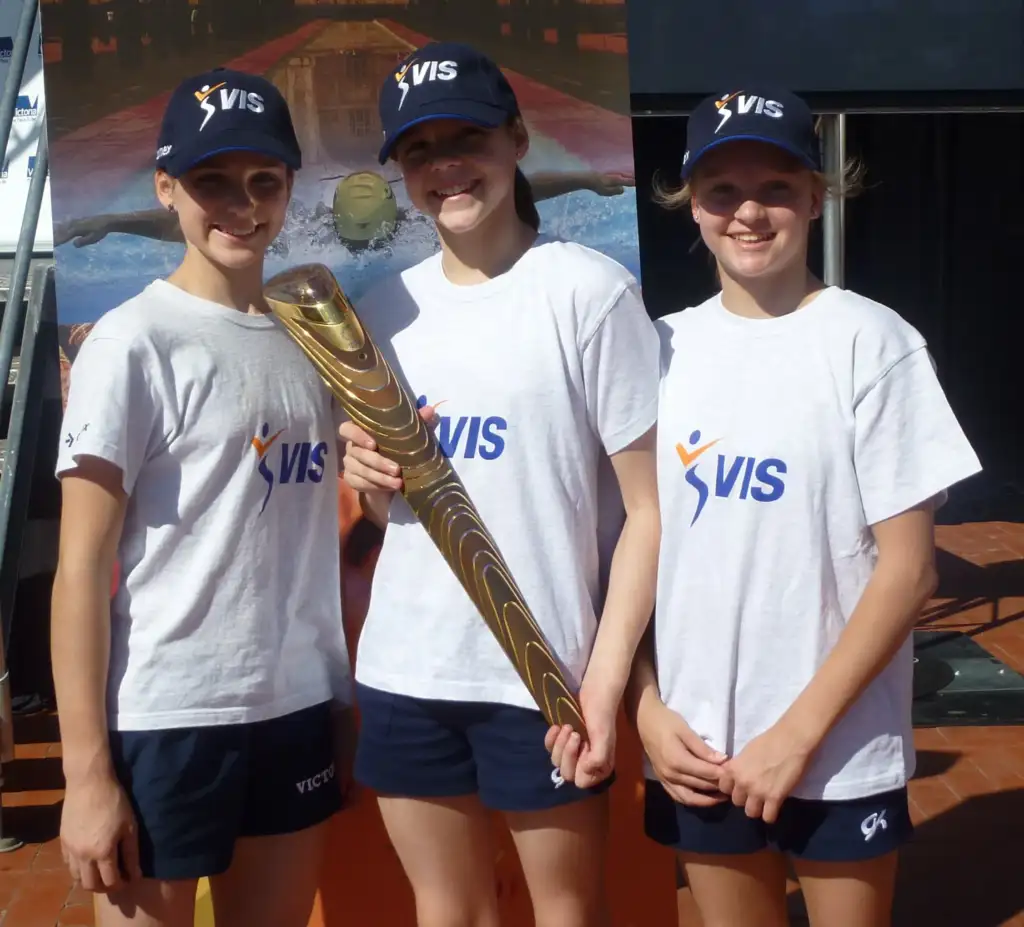 Three young VIS female athletes, all wearing white tees with the VIS logo on the front and navy VIS branded caps. They are holding a Commonwealth Games torch. 