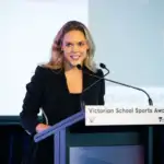 A female in her early thirties with long wavy blonde hair, wears a black blazer. She is standing behind a lectern, presenting.