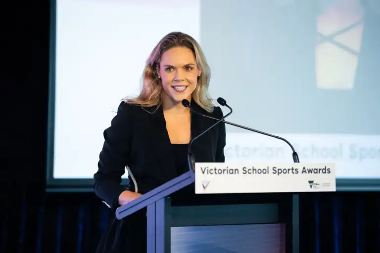 A female in her early thirties with long wavy blonde hair, wears a black blazer. She is standing behind a lectern, presenting.