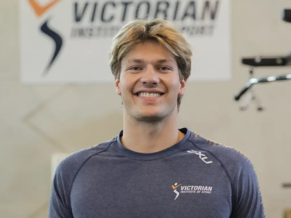 A headshot of a male athlete with short blonde hair. He stands in front of a blurred background with gym equipment and a sign reading Victorian Institute of Sport.