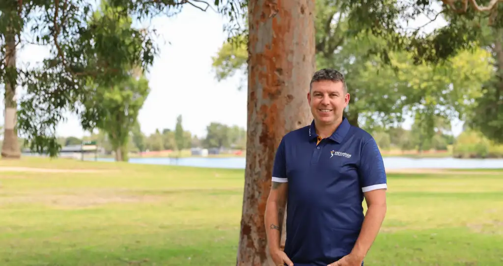 A picture of a man in his forties wearing a VIS polo, standing in front of a tree with a lake in the background, smiling to the camera.