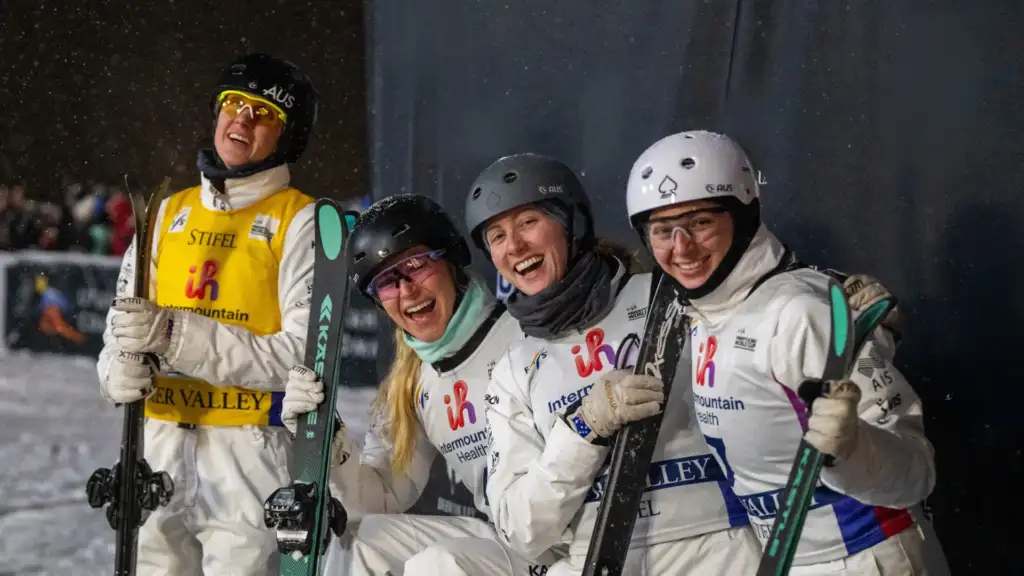 Four VIS aerial skiing athletes wearing white outwear and helmets, holding skis, all smiling to the camera.