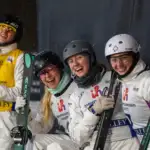 Four VIS aerial skiing athletes wearing white outwear and helmets, holding skis, all smiling to the camera.