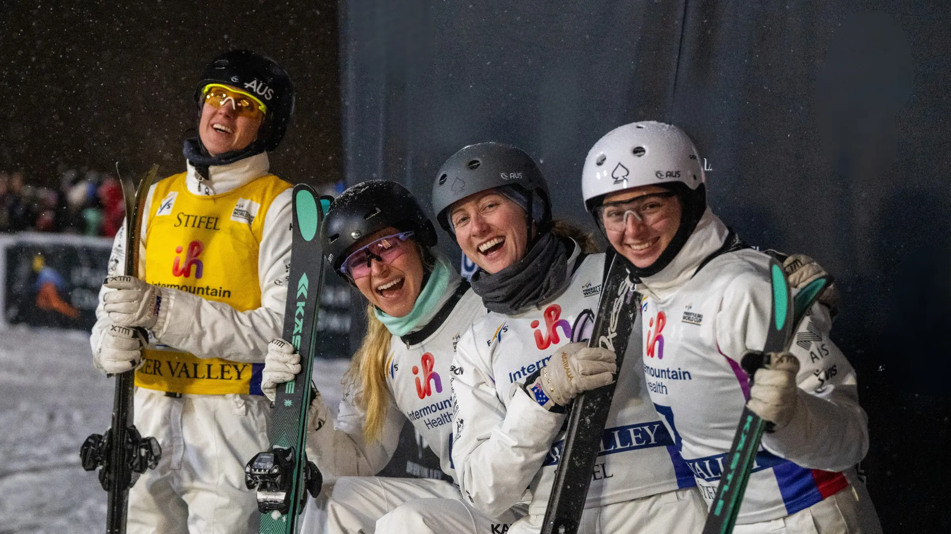 Four VIS aerial skiing athletes wearing white outwear and helmets, holding skis, all smiling to the camera.