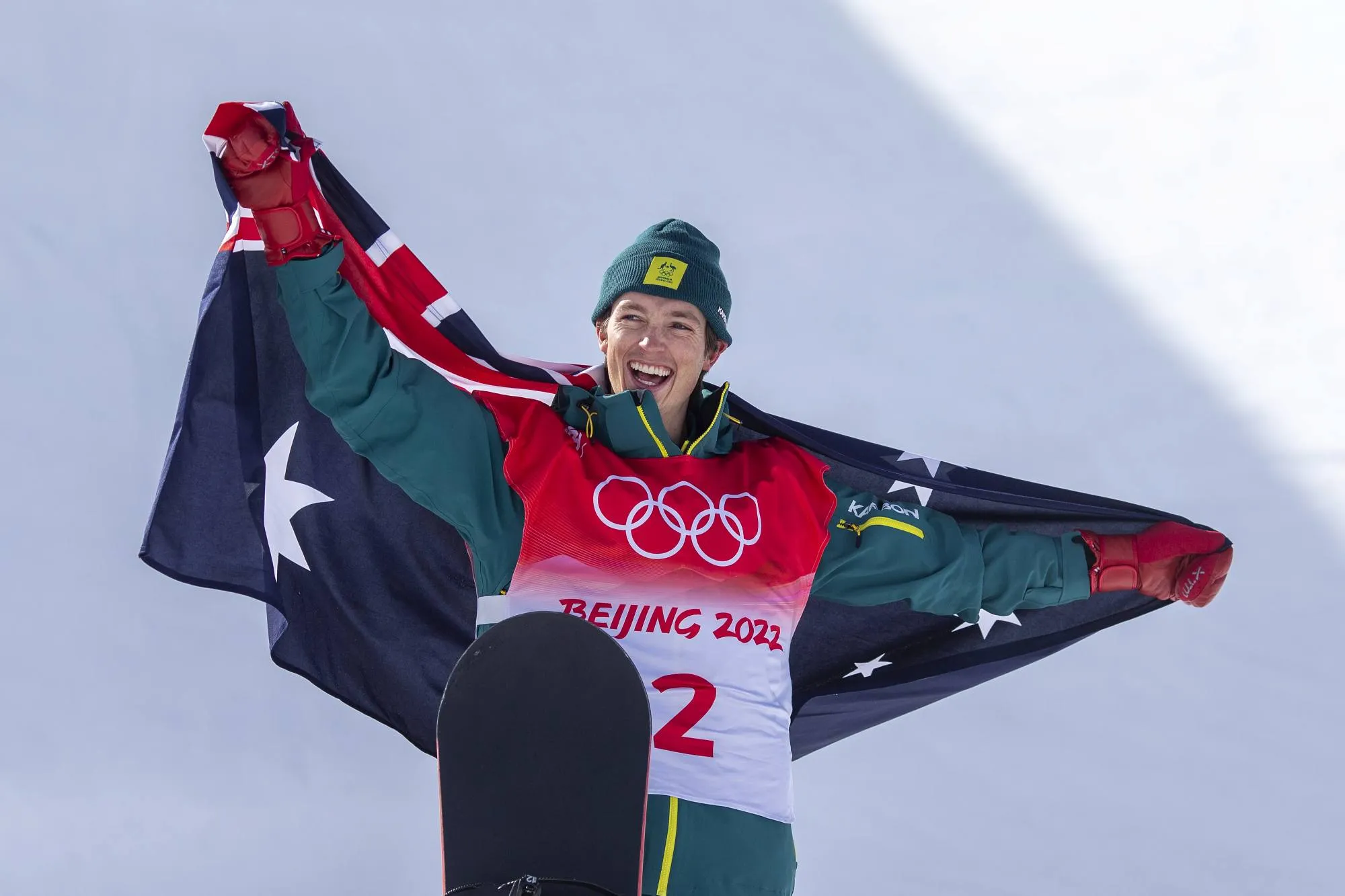 A male athlete in his late twenties wears a green Australian Olympic Team snow jacket and a 2022 Beijing Winter Olympic Games bib, he's smiling and holding up an Australian flag.