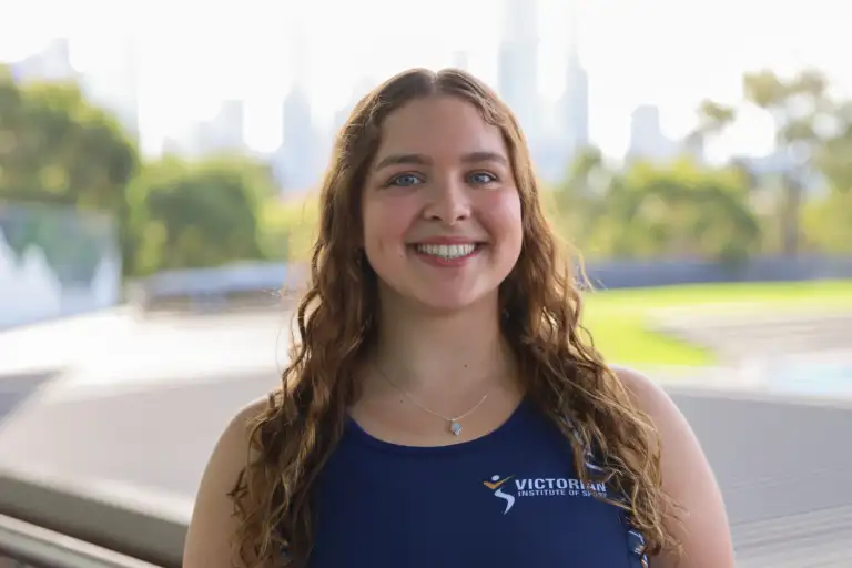 A headshot of a female athlete with long curly brown hair which sits over her shoulders. She wears a navy singlet and a blurred cityscape is in the background.