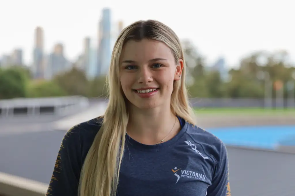 A headshot of a female athlete with long straight blonde hair which sits over her left shoulder. She wears a navy tee and a blurred cityscape is in the background.