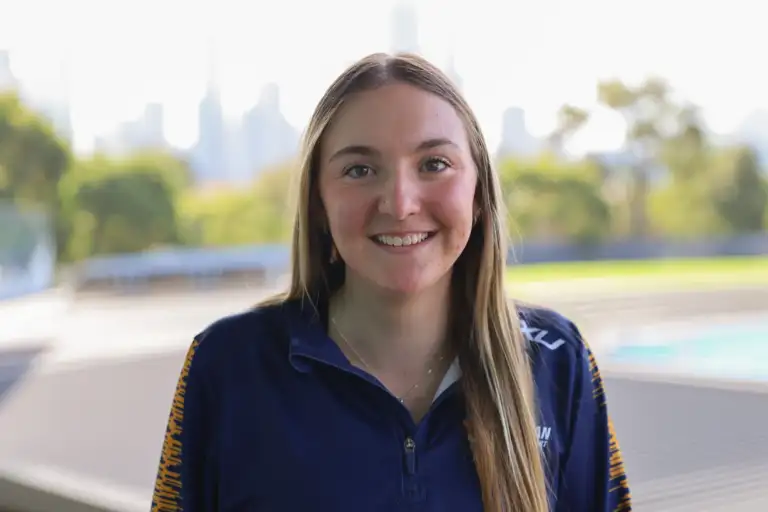 A headshot of a female athlete with long straight blonde hair which sits over her right shoulder. She wears a navy quarter zip top and a blurred cityscape is in the background.