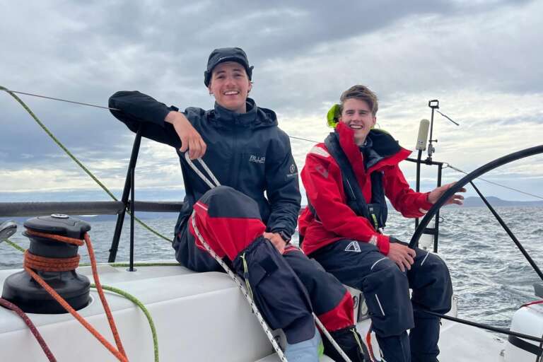 A photo of two men in their twenties sitting on the side of a sailing boat at sea. One wears a black jacket and cap, the other a red jacket with short brown hair. Both are smiling to the camera.