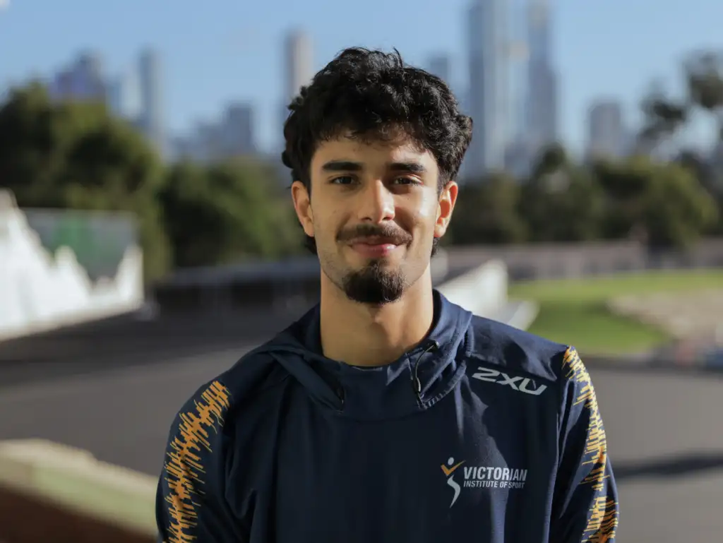 A headshot of a male athlete with short black wavy hair and short facial hair. He wears a navy quarter zip top with the VIS logo on the left chest and stands in front of a blurred cityscape in the background