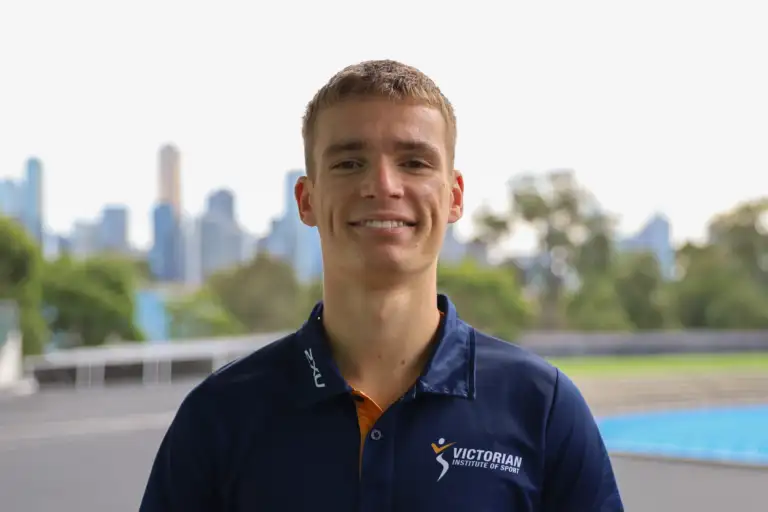 A headshot of a male athlete with short blonde hair. He wears a navy polo with the VIS logo on the left chest and stands in front of a blurred cityscape in the background.