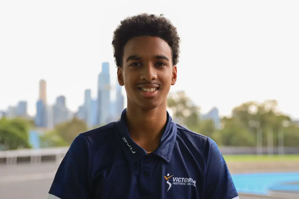 A headshot of a male athlete with short black curly hair. He wears a navy polo with the VIS logo on the left chest and stands in front of a blurred cityscape in the background