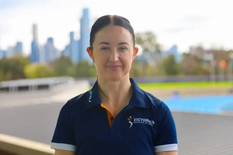 A headshot of a female athlete with dark brown hair tied back. She's wearing a navy VIS branded polo with a blurred cityscape behind her.