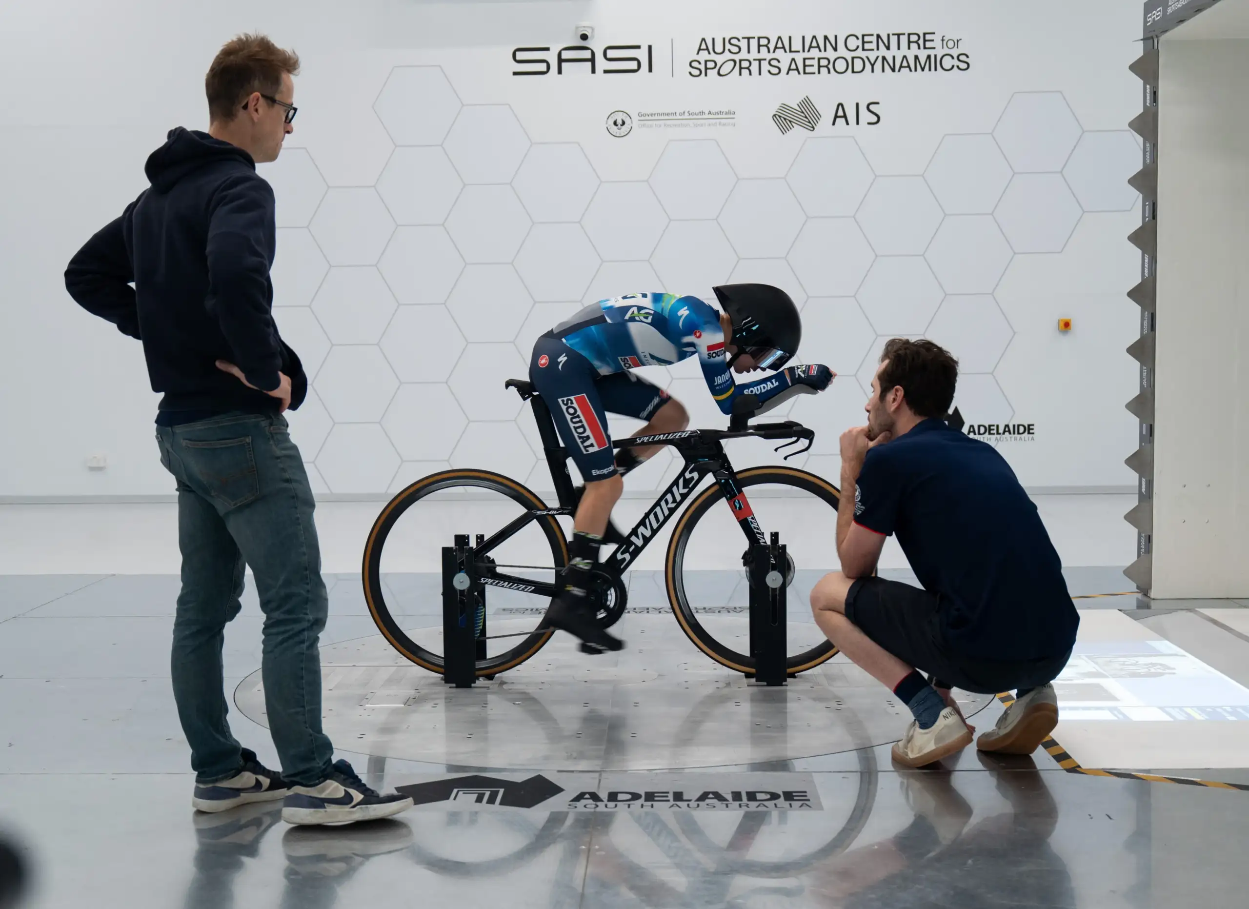 A female athlete is on a stationary bike with two aerodynamic technicians watching. She pedals in a white room with a forward wind simulator.