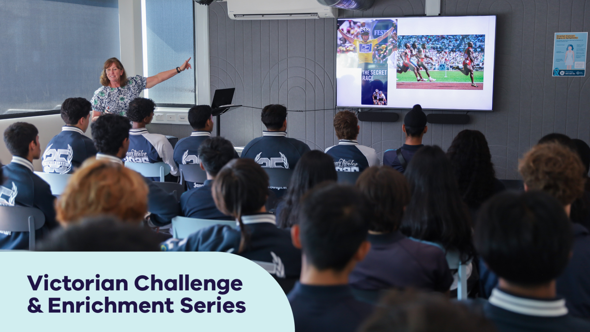 A room of twenties high school students, listening to a female in her fifties present at the front of a room. She has long brown curly hair and is pointing to a powerpoint presentation on screen.
