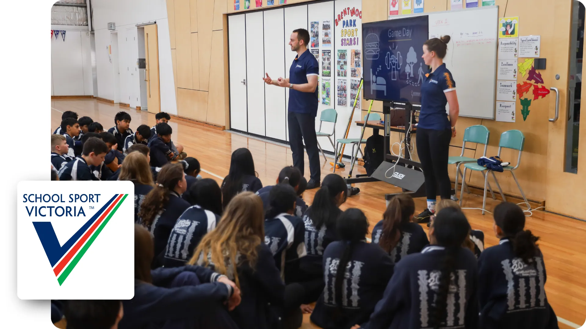 Two VIS athletes, one male and one female, stand in front of a room of primary school aged children, doing a presentation.