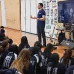 Two athletes, one male and one female in their twenties and thirties are presenting to a group of school children who sit on a gym floor engaged.