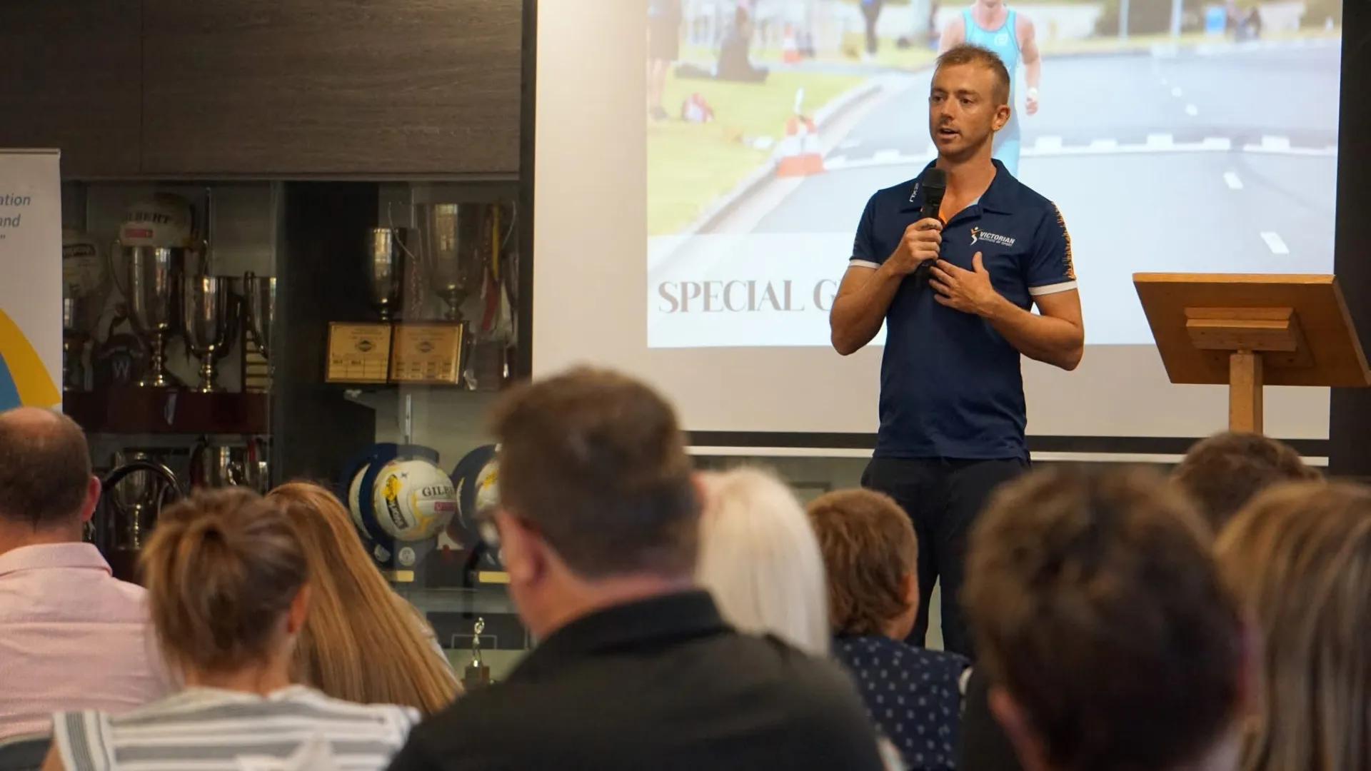 A photo of a male with short blonde hair in his twenties, public speaking to a room of adults.