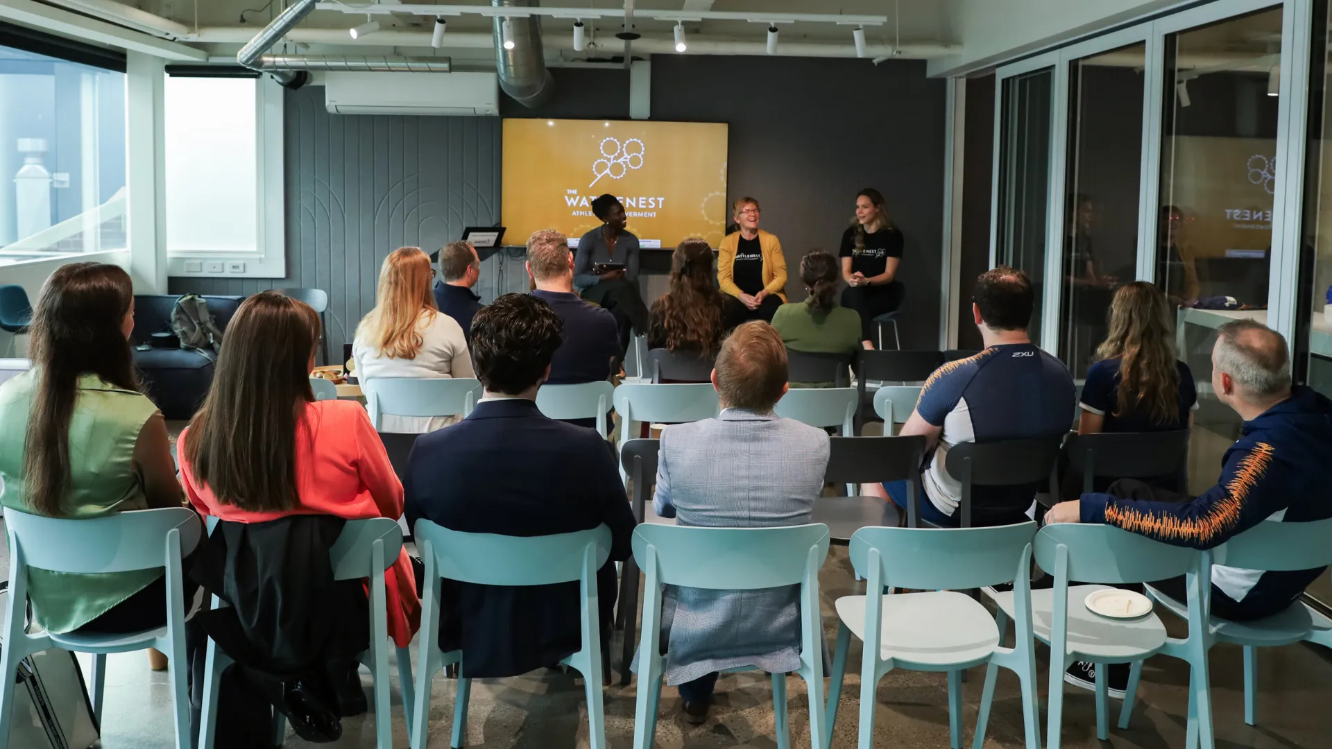 A photo of a medium sized room full of people in seats, listening to a panel of three women sitting on stools at the front of the room.
