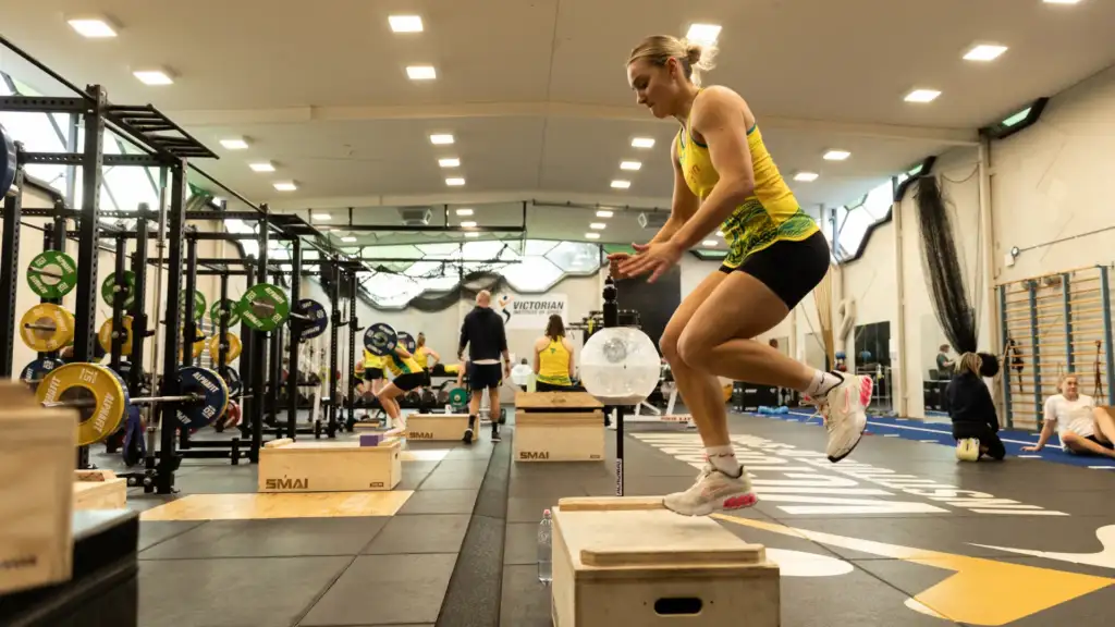 A photo of a female athlete in her twenties with blonde hair, wearing Australian diamonds uniform jumping onto a box in a gymnastium. In the background are more athletes in the same uniform working out in the gym.