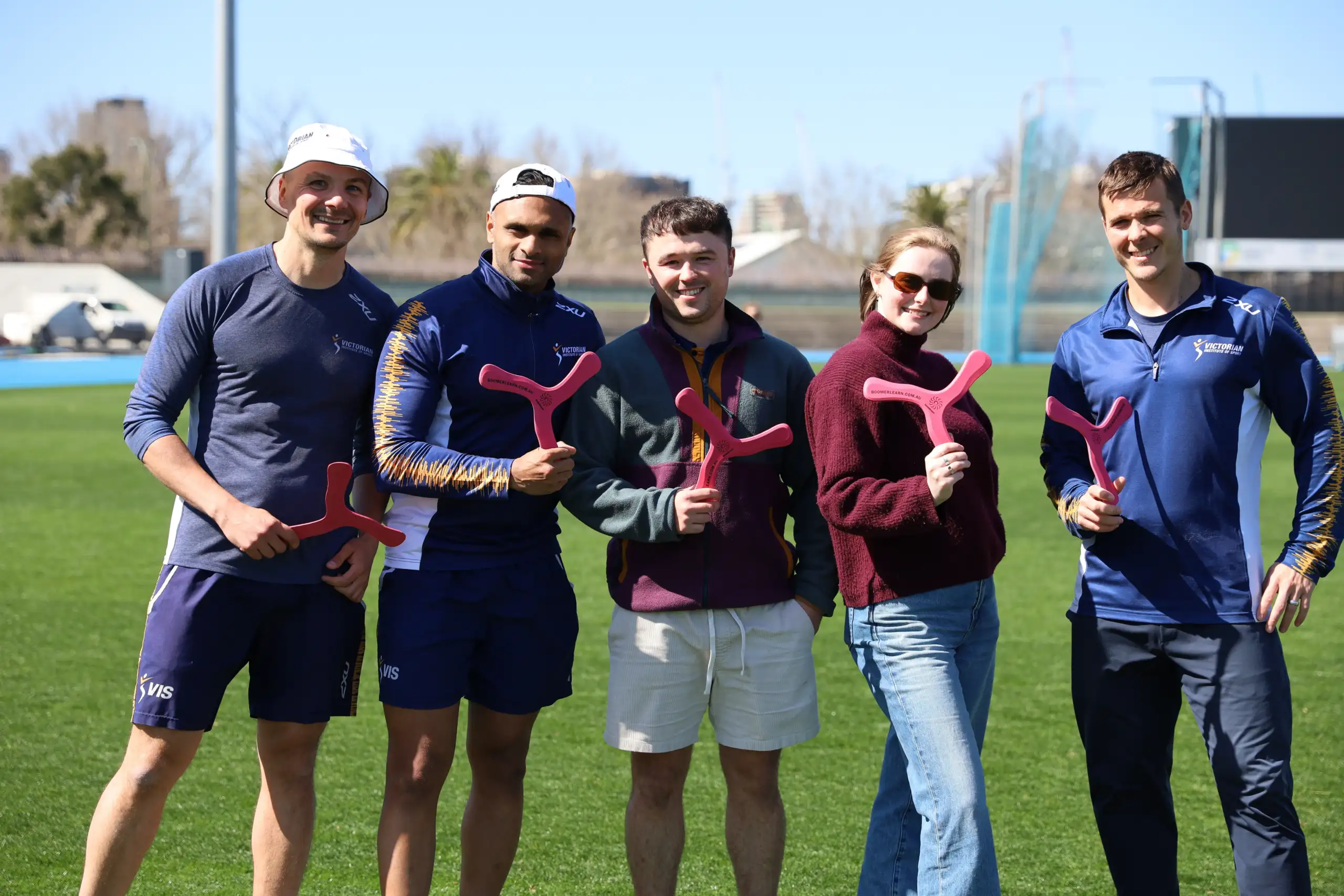 A photo of five adults, four males and one female, standing on a green pitch holding up pink three-pronged boomerangs.