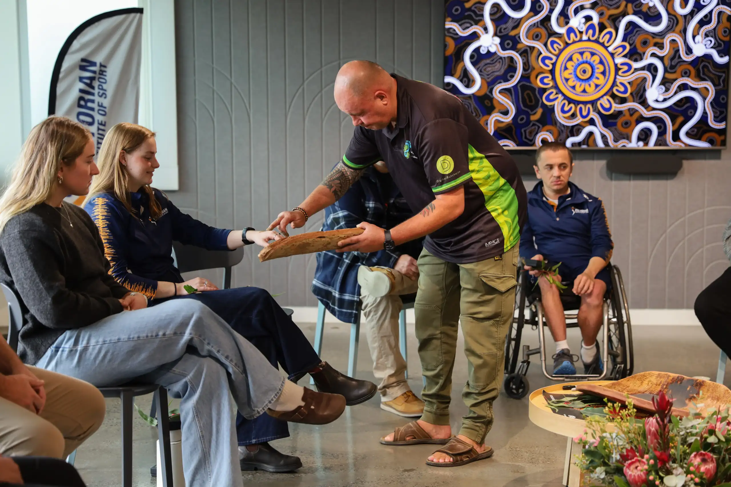 A man in his fifties wearing green utility pants and a black and green polo, is painting ochre onto a female's hand.