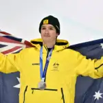 A male in his thirties wearing a yellow Australian Olympic Team snow jacket and holding up an Australian flag behind him, with a silver medal around his neck.