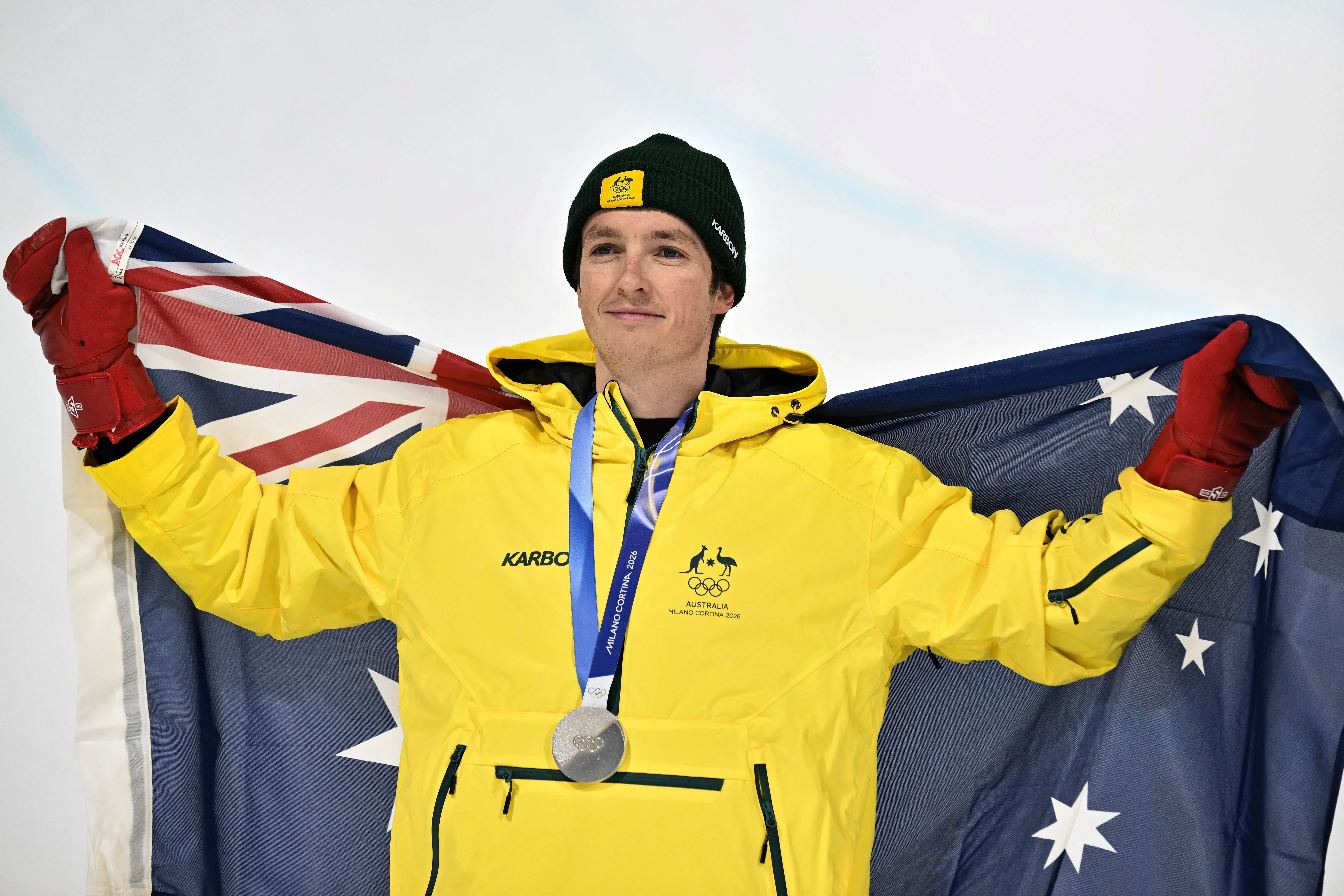 A male in his thirties wearing a yellow Australian Olympic Team snow jacket and holding up an Australian flag behind him, with a silver medal around his neck.