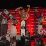 Three male snowboarding athletes stand on a podium, the middle being VIS scholarship athlete, Scotty James. He wears a brown jacket, has short brown hair and has his arms up in triumph.